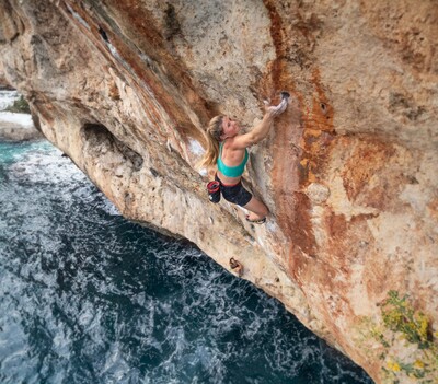 A woman climbs on the steep sand-colored rock directly above the sea.