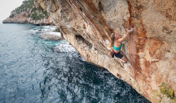 A woman climbs on the steep sand-colored rock directly above the sea.